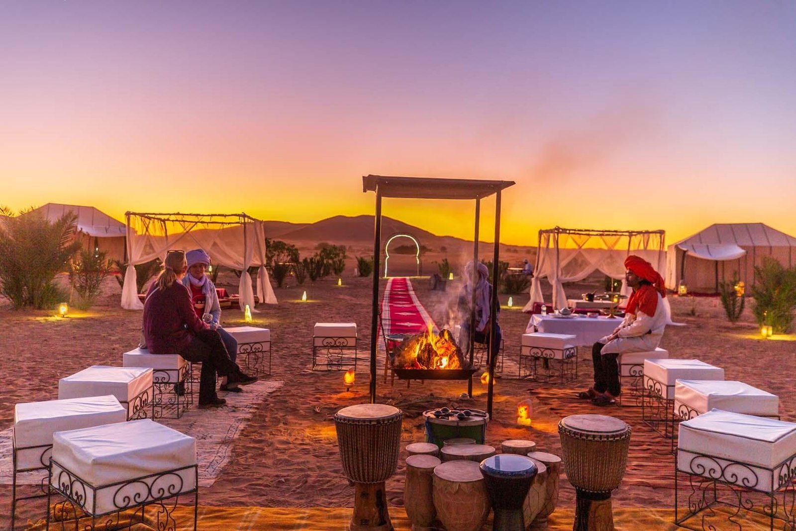Campamento de lujo en el desierto del Sáhara al atardecer, con fuego, tambores y salones bajo toldos en un viaje de lujo a Marruecos